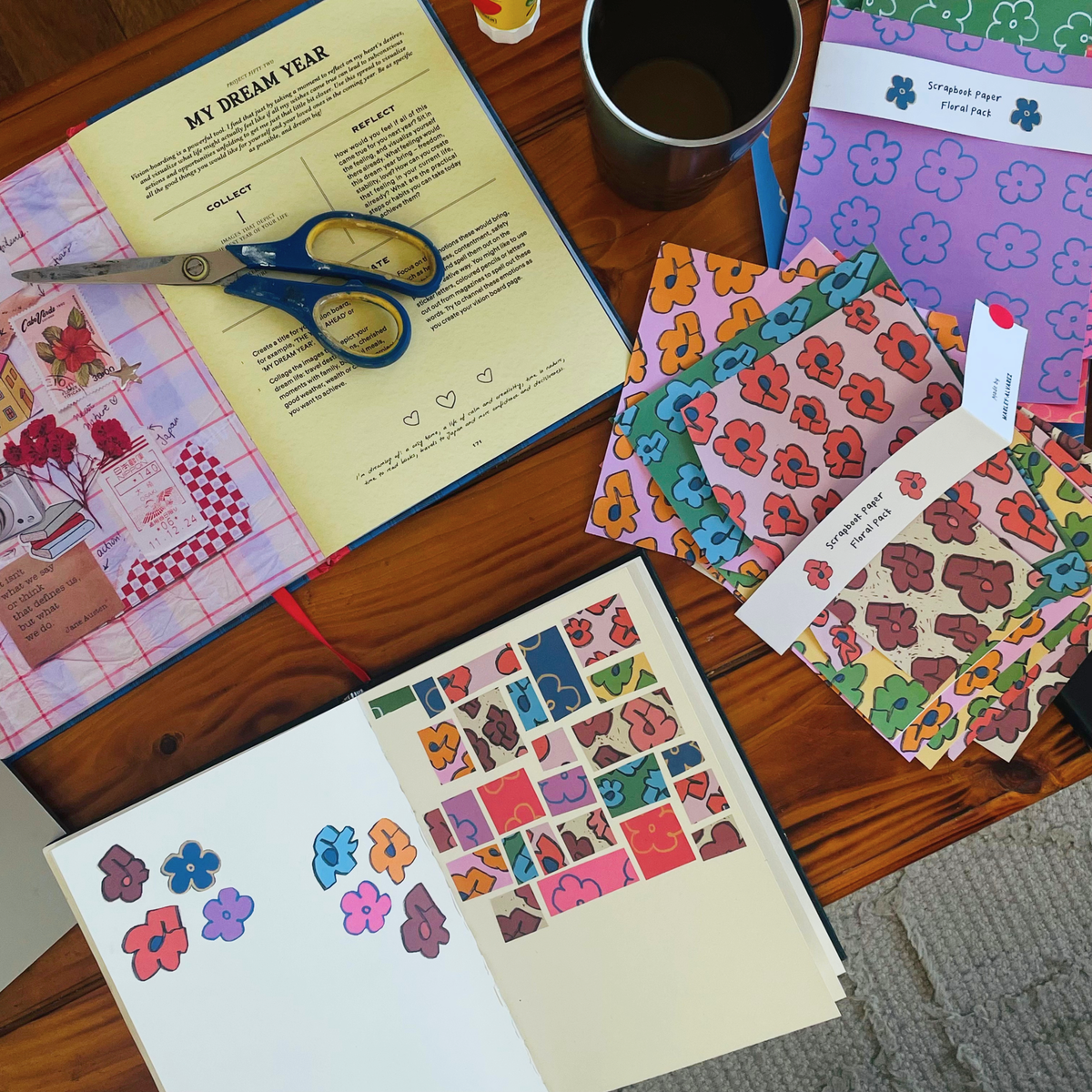 A styled flat-lay of a handmade journaling and scrapbooking kit on a wooden table, featuring patterned paper sheets, a decorated journal page, a pair of blue-handled scissors, and an open book with 'My Dream Year' instructions.