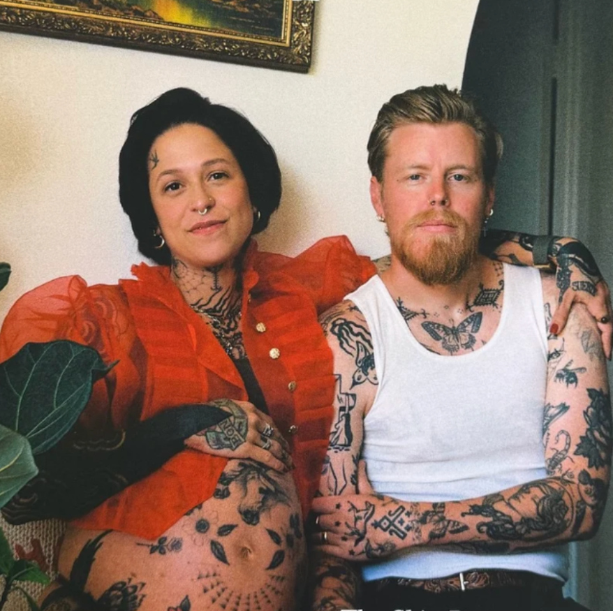 Two people with tattoos sitting together in a room with a plant and framed picture.